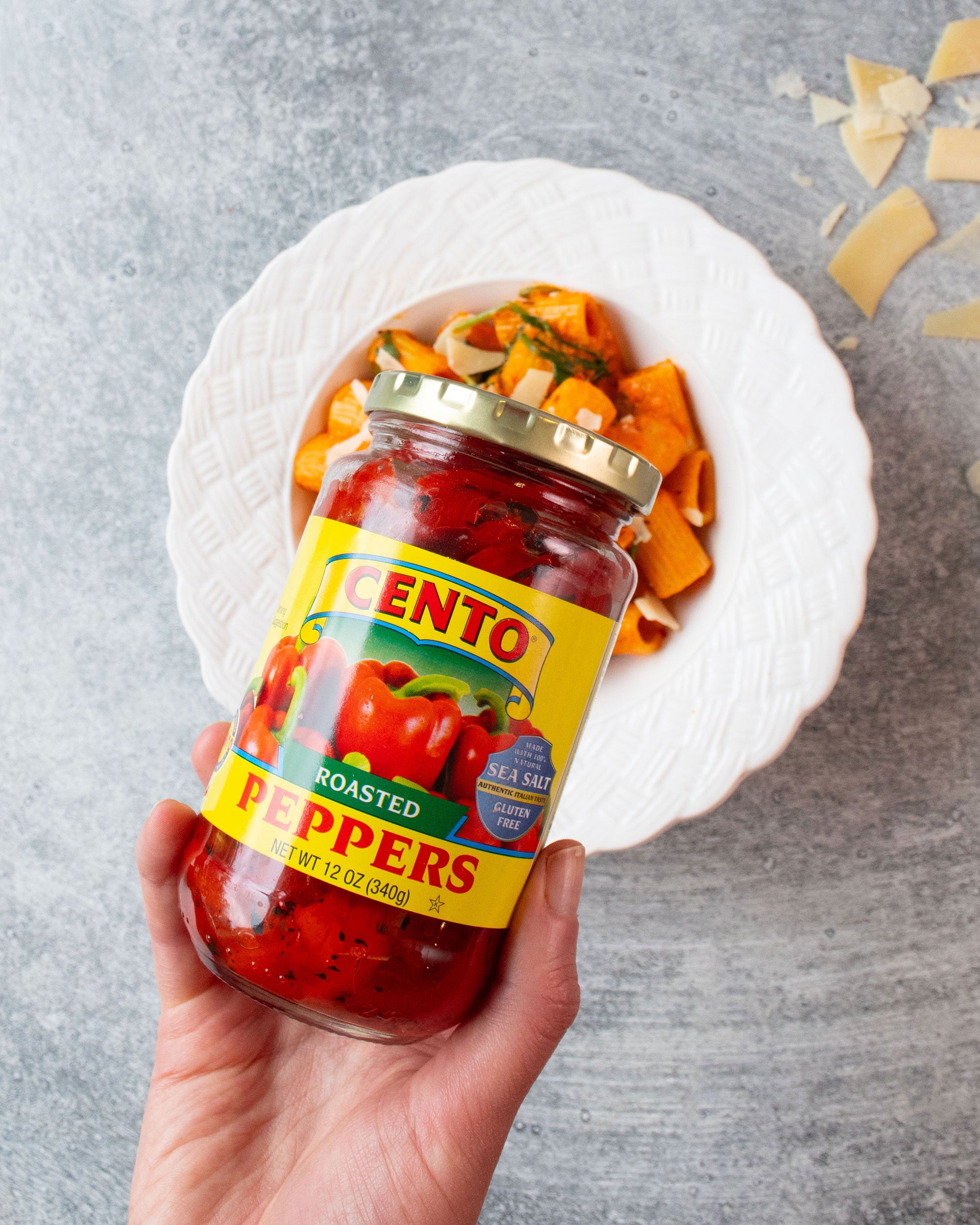 Hand holding a jar of Cento roasted peppers with a bowl of pasta in the background.