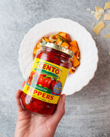 Hand holding a jar of Cento roasted peppers with a bowl of pasta in the background. thumbnail